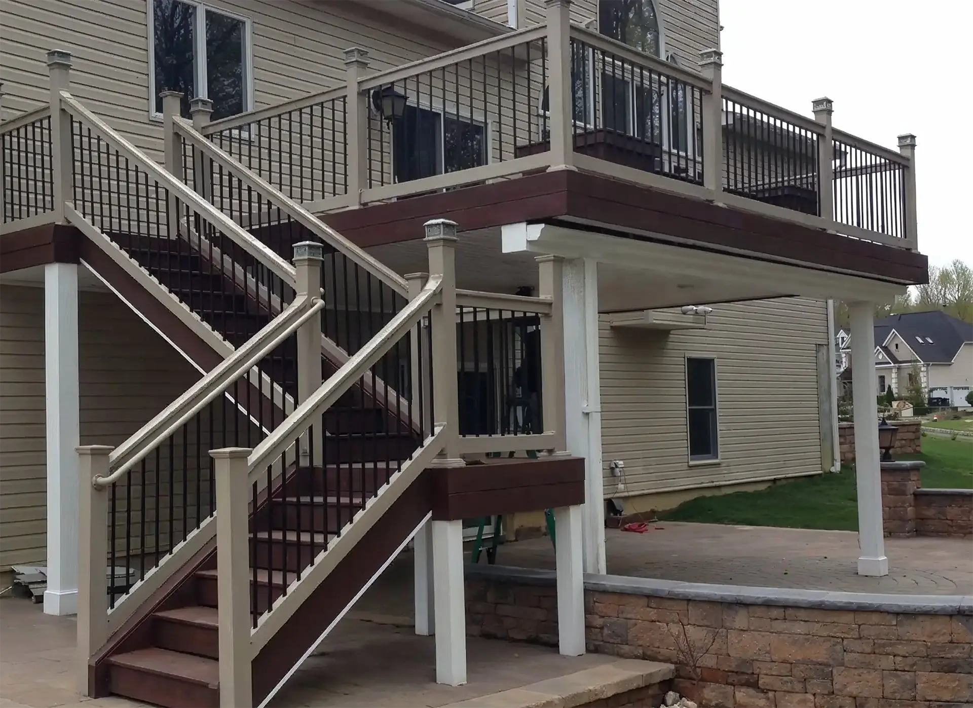 An elevated, two-story composite deck addition showcasing custom deck building craftsmanship. Features include a two-tone design with dark brown fascia trim, tan railings with black metal balusters, integrated post cap lighting, a wide staircase with central safety handrails, and heavy-duty white support columns over a stone paver patio.